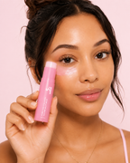 A woman holding a pink cosmetic stick product next to her cheek, showing a glossy highlighter or bronzer effect. The product packaging is light pink with white text, and the background is a solid light pink.